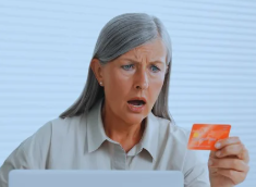 A woman with gray hair, wearing a beige blouse, sits at a table with a white laptop in front of her. She holds a credit card in her left hand and looks shocked and worried. The image represents online fraud, possibly due to stolen credit card information.