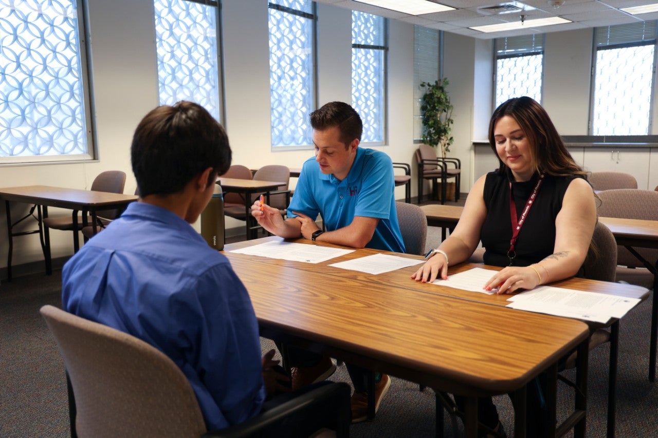 Three people sit around a table. One person is facing the other two as though for a meeting. The remaining two people are looking down at pieces of paper.
