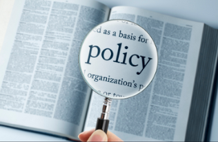 An open book is lying on a table. A large magnifying glass is positioned above it, making the word "policy" appear bigger and bolder. The image shows a close examination of rules and guidelines.