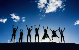 Seven people are jumping  together on an elevated surface. The background shows a sunset with soft clouds. Bright light appears around their feet, emphasizing movement and excitement. The image shows excitement and happiness.