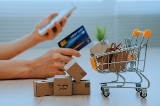 The image shows a woman's hands. She holds a cellphone in her left hand and a credit card in her right. To the right of her hand, there are four small boxes, one of which has the words "Online Shopping" written on it. Next to the boxes is a tiny shopping cart, like the ones in grocery stores, filled with small paper shopping bags. The image represents online shopping.