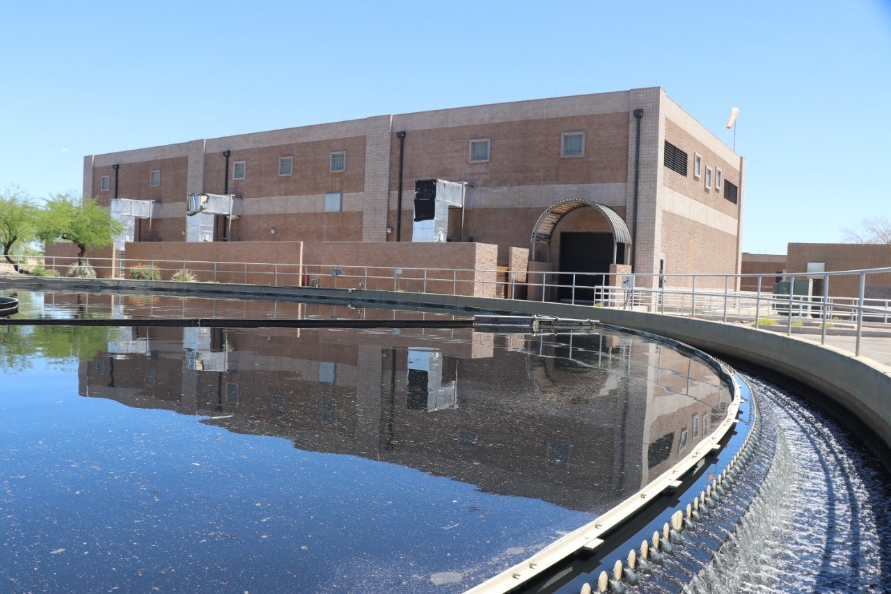 This image shows the Phoenix Water Services 91st Avenue Wastewater Treatment Plant. A circular water treatment basin with flowing water is in the foreground, reflecting the facility's main building in the background. The building is constructed of brown brick and features multiple small windows and ventilation structures. There are railings and surrounding greenery, including trees and desert landscaping, under a clear blue sky.